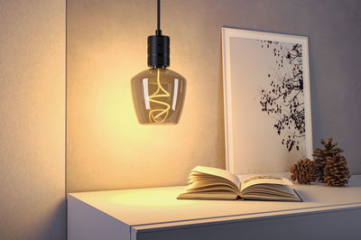 Hanging black metal lamp with warm light above white table with book and decorative pine cones.