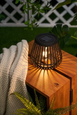 Black metal solar table lamp with warm light on wooden bench in garden setting with plants.