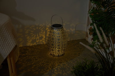 Dark grey metal lantern with intricate pattern casting warm light and decorative shadows indoors.