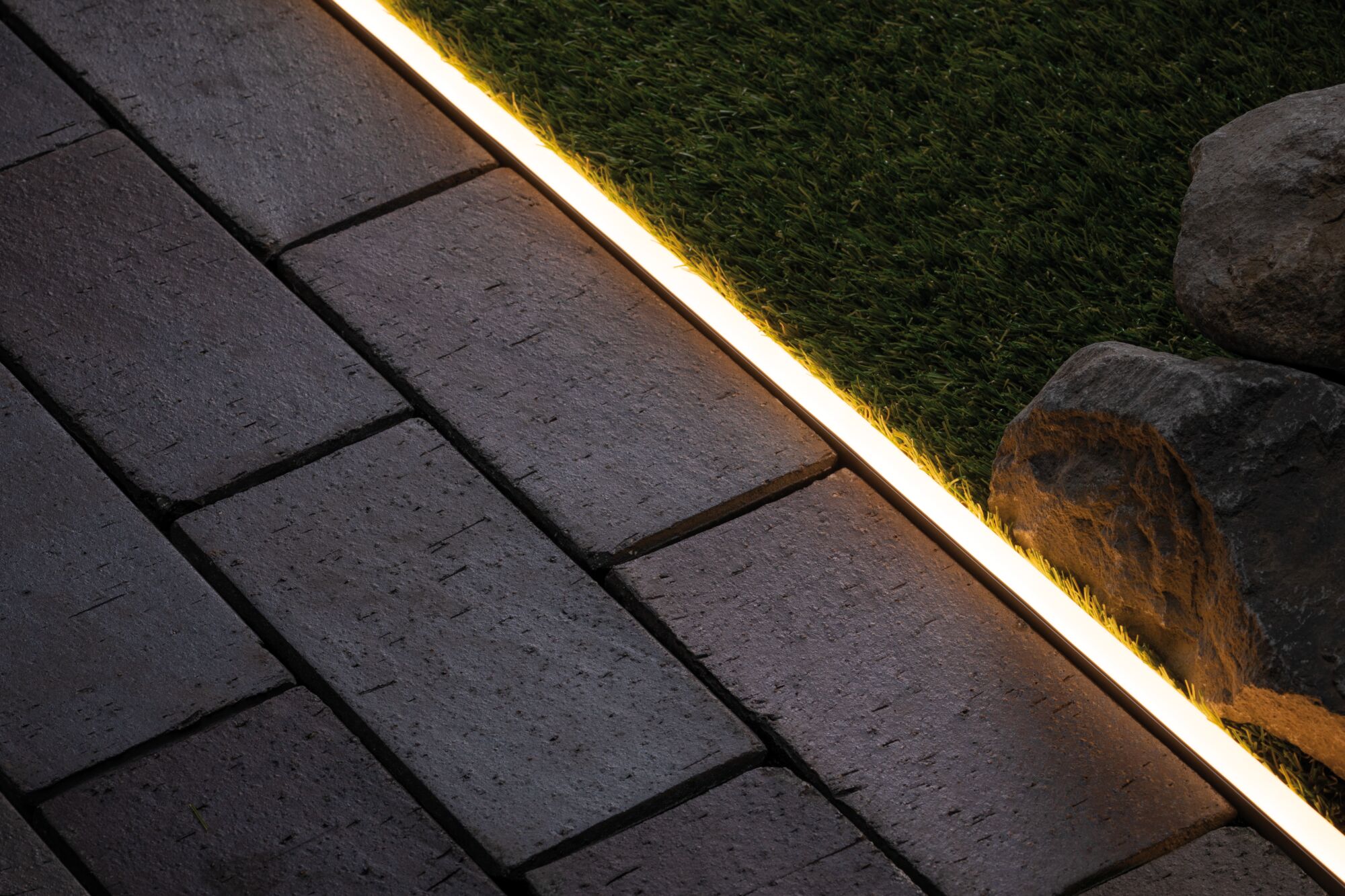 Warmweißes LED-Bandlicht zwischen grauen Pflastersteinen und grünem Rasen für Außenbeleuchtung.