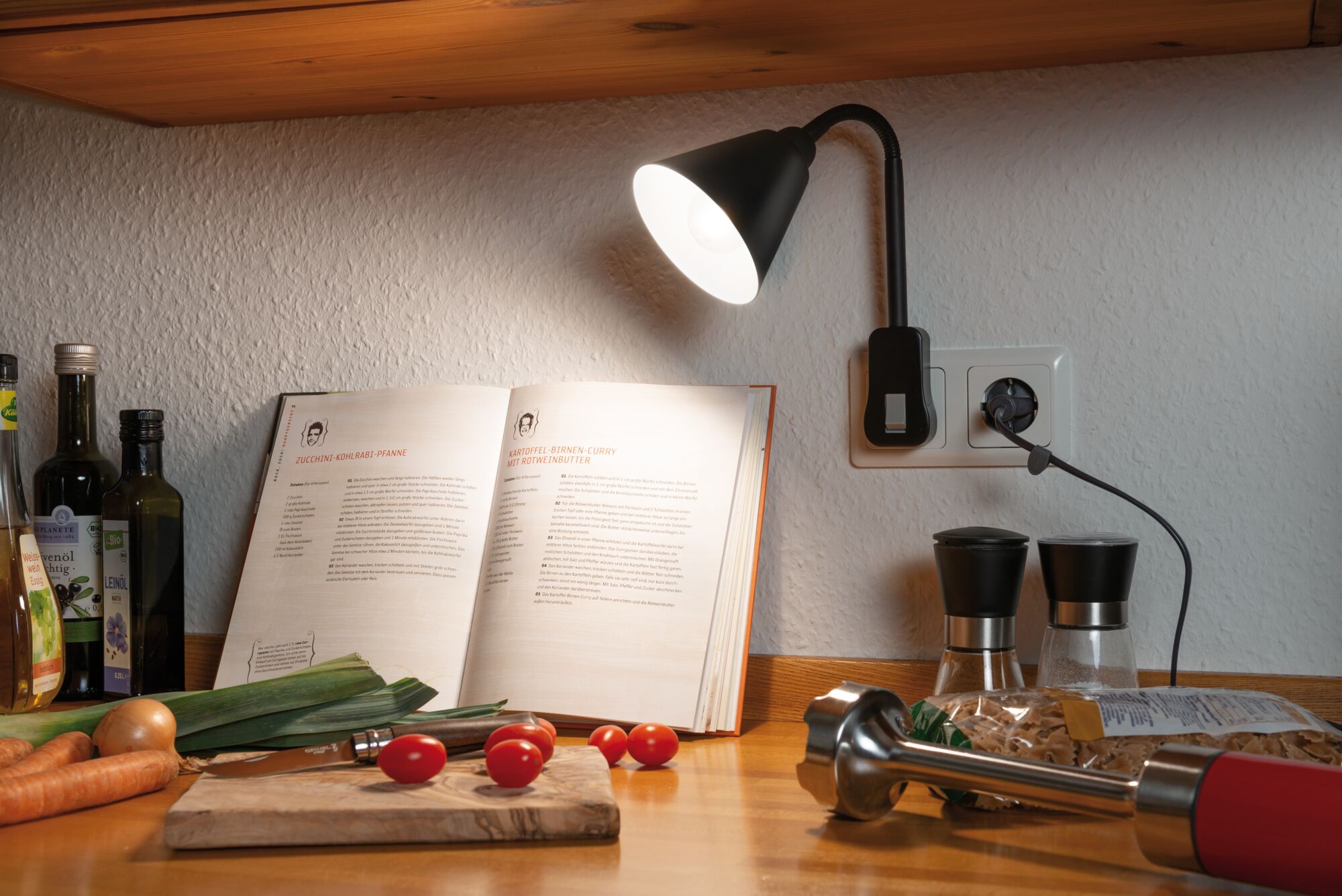 Lampe LED noire branchée sur prise murale de cuisine éclairant un livre de recettes et légumes frais.