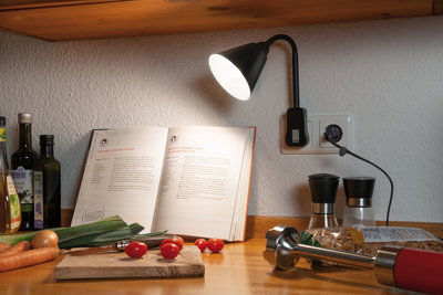 Lampe LED noire branchée sur prise murale de cuisine éclairant un livre de recettes et légumes frais.