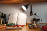 Black LED plug-in lamp on kitchen wall lighting cookbook and fresh vegetables on wooden countertop.