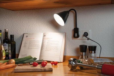 Black LED plug-in lamp on kitchen wall lighting cookbook and fresh vegetables on wooden countertop.