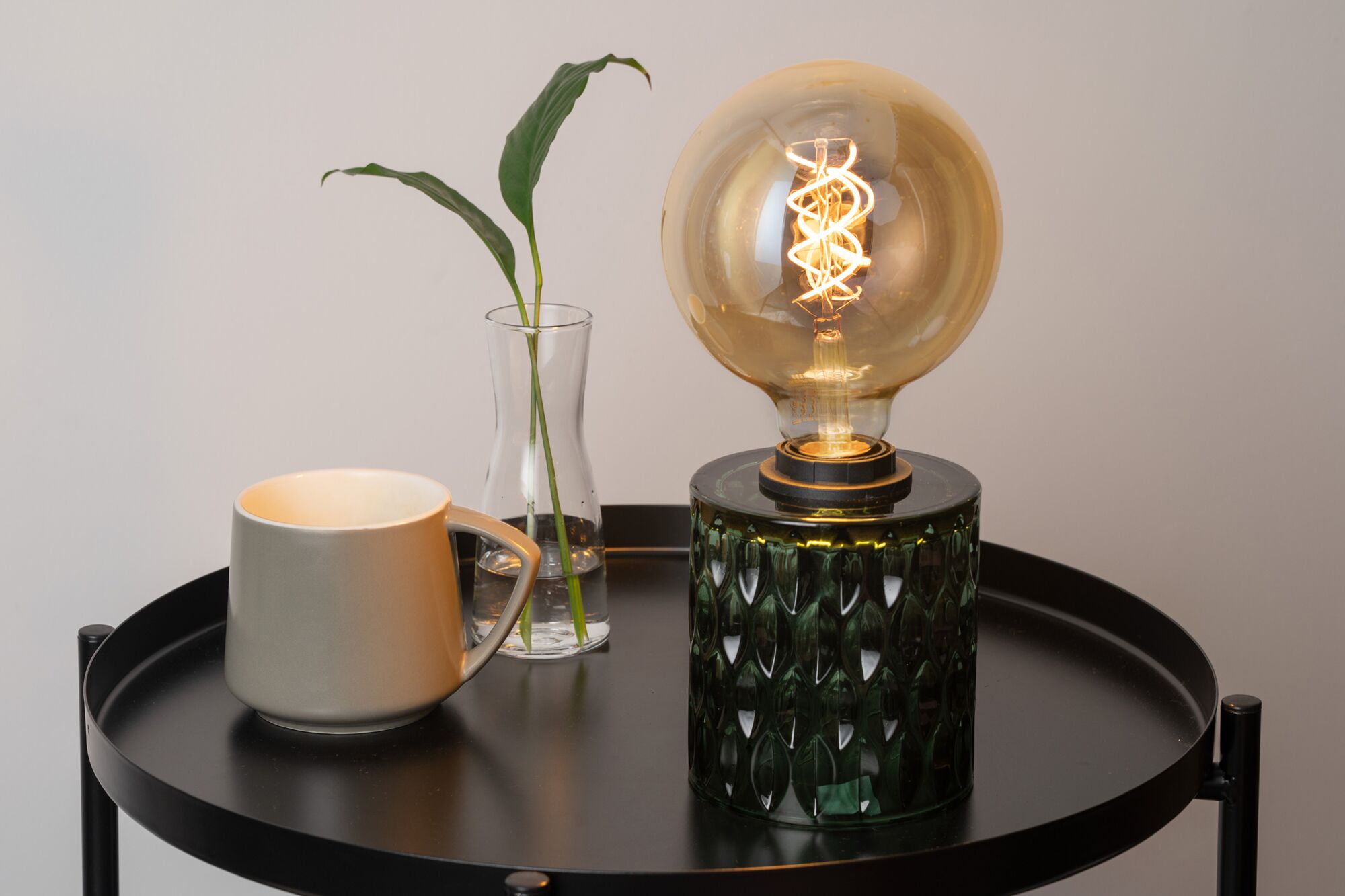 Grøn glasbordlampe med dekorativ pære på sort metalbord ved siden af grå kop og glasvase
