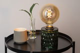 Grøn glasbordlampe med dekorativ pære på sort metalbord ved siden af grå kop og glasvase