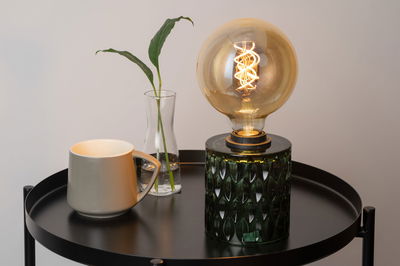 Grøn glasbordlampe med dekorativ pære på sort metalbord ved siden af grå kop og glasvase
