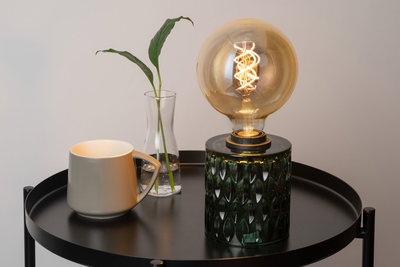 Grüne Glas-Tischlampe mit dekorativer Glühbirne auf schwarzem Metalltisch neben grauer Tasse und Glasvase