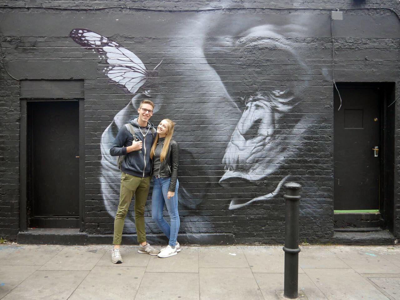 Junges Paar vor schwarzer Wand mit Graffiti eines Schmetterlings und Gesichtsprofil in urbaner Straße