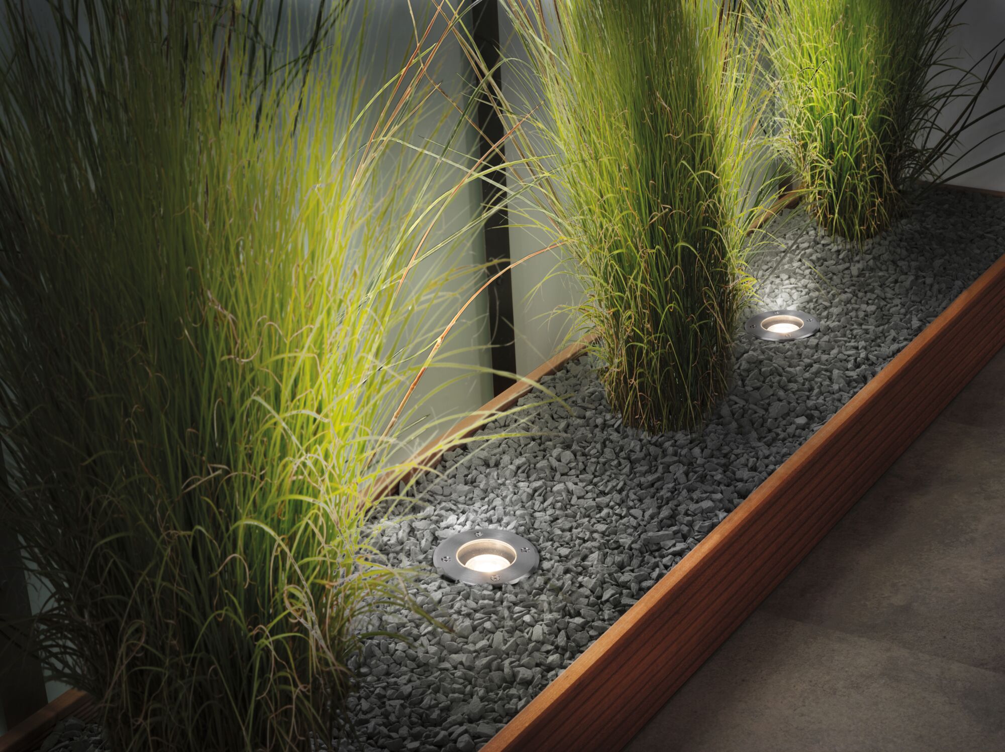 Recessed stainless steel LED floor lights illuminating green plants in gravel bed with wooden frame