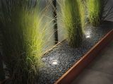 Recessed stainless steel LED floor lights illuminating green plants in gravel bed with wooden frame