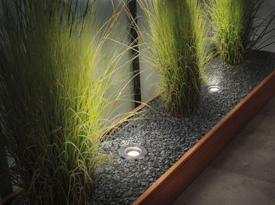 Recessed stainless steel LED floor lights illuminating green plants in gravel bed with wooden frame