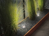 Recessed stainless steel LED floor lights illuminating green plants in gravel bed with wooden frame