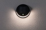 Round black solar wall light with motion sensor and warm white LED on textured grey wall