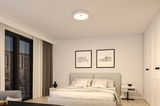Rundes weißes Deckenlicht aus Kunststoff in modernem Schlafzimmer mit grauem Bett und weißen Wänden.