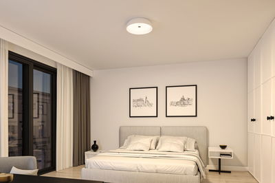 Rundes weißes Deckenlicht aus Kunststoff in modernem Schlafzimmer mit grauem Bett und weißen Wänden.