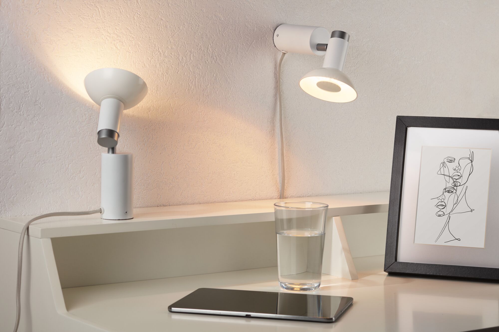 Zwei weiße LED-Wandleuchten aus Metall an strukturierter Wand mit Glas Wasser und Tablet auf Tisch