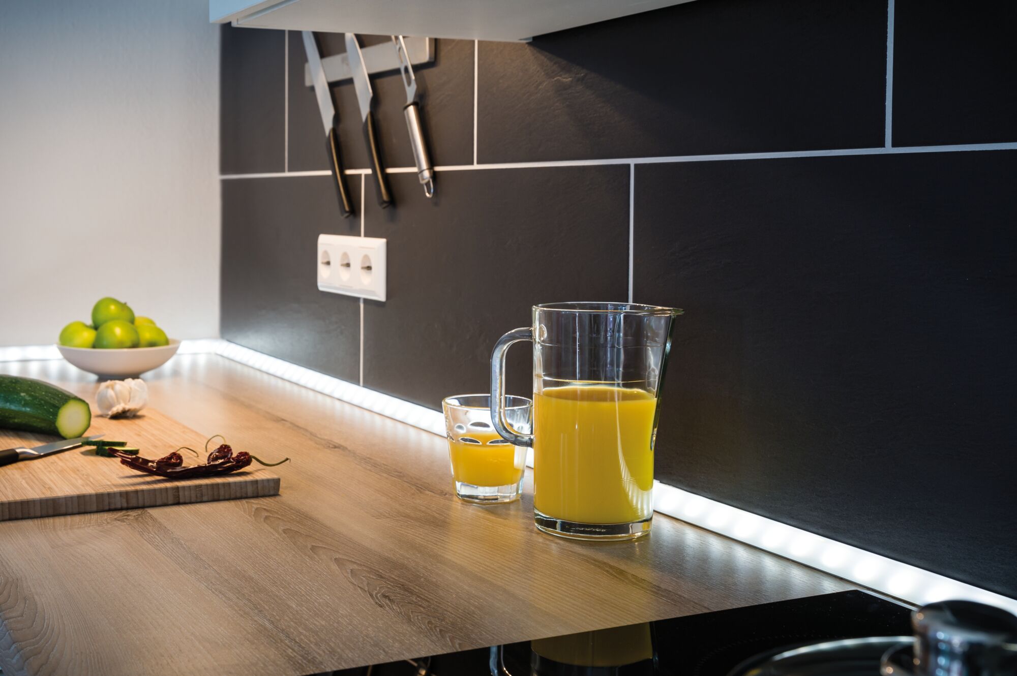 Modern kitchen countertop with LED under-cabinet lighting, glass jug of orange juice and black tile backsplash