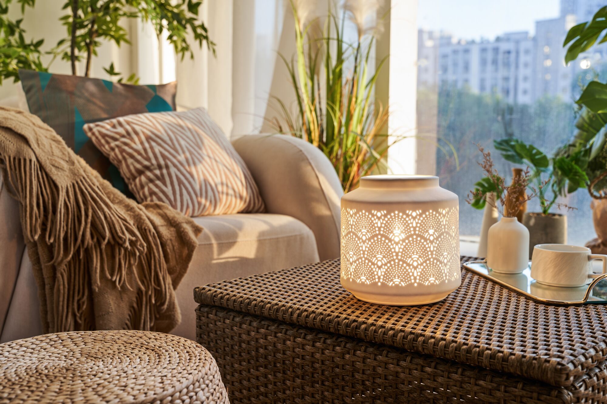 Beleuchtete weiße Keramiklampe mit filigranem Muster auf geflochtenem Tisch im Wohnzimmer mit Kissen