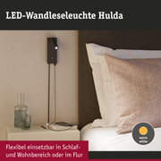 Schwarze LED-Wandleuchte Hulda mit flexiblem Lesearm und warmweißem Licht neben Bett und Nachttisch