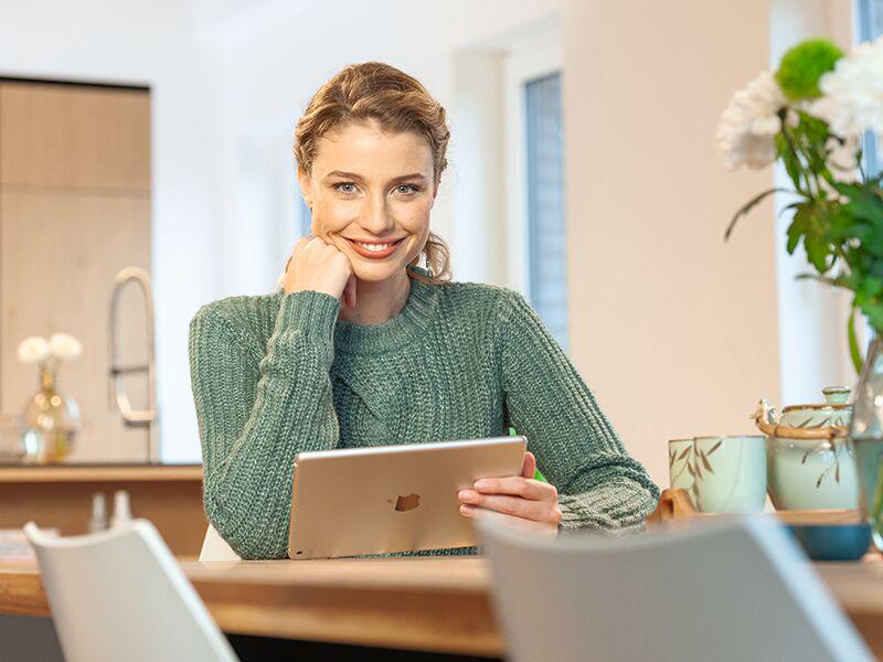 Frau mit grünem Strickpullover sitzt am Holztisch und nutzt ein Tablet in hellem Wohnraum mit moderner Einrichtung
