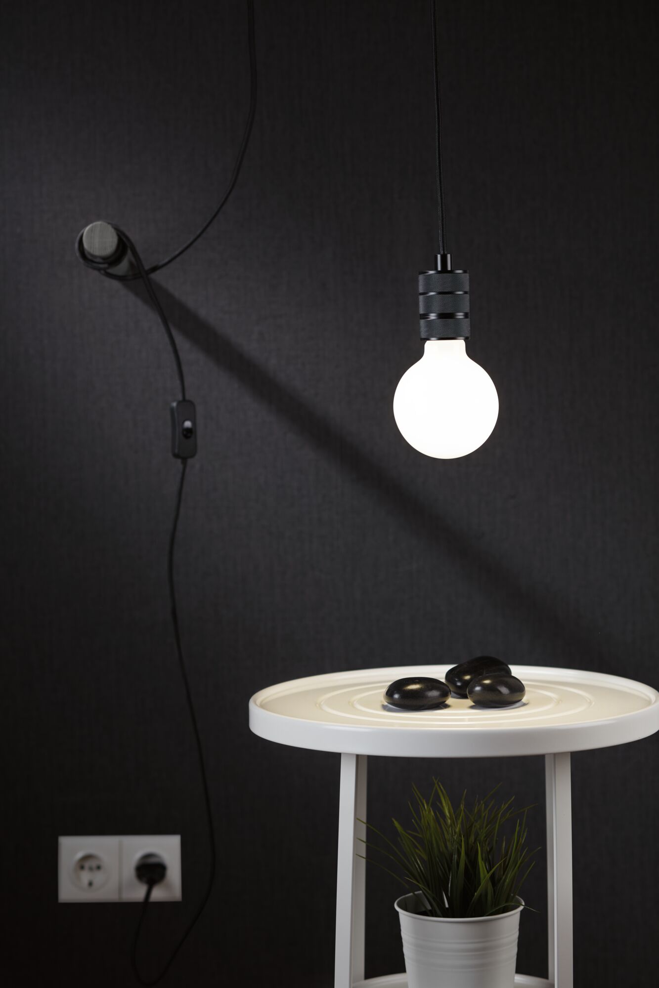 Ampoule LED blanche suspendue avec câble noir au-dessus d'une table d'appoint blanche avec plante et pierres décoratives