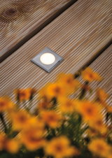 Quadratischer LED-Bodeneinbaustrahler aus gebürstetem Edelstahl auf Holzterrasse mit Blumen.