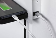 Nahaufnahme eines Smartphones mit grünem Ladebalken an einem silbernen, geflochtenen Ladekabel an weißer Wand.