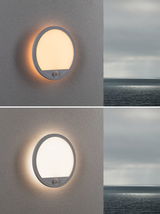 Runde weiße Außenwandleuchte mit Bewegungsmelder und warmweißem Licht an grauer Wand mit Meerblick