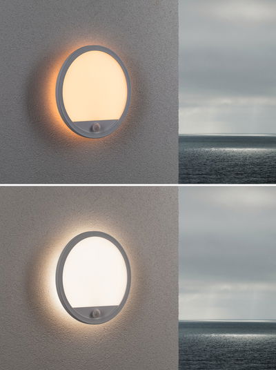 Runde weiße Außenwandleuchte mit Bewegungsmelder und warmweißem Licht an grauer Wand mit Meerblick
