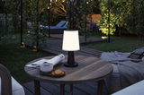 Black outdoor table lamp with white shade on wooden table in garden setting with ambient lighting and lounge.