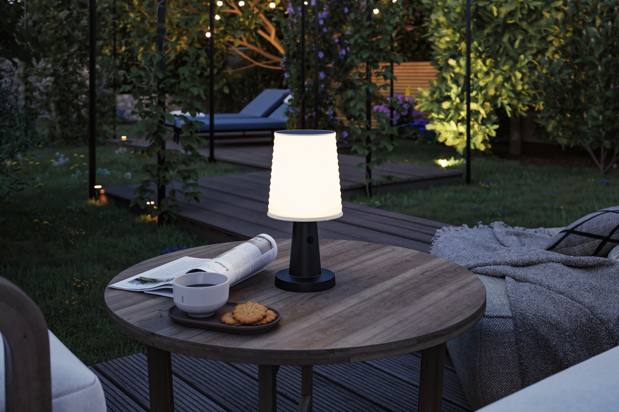 Schwarze Outdoor-Tischlampe mit weißem Lampenschirm auf Holztisch im Garten mit Beleuchtung und Lounge.