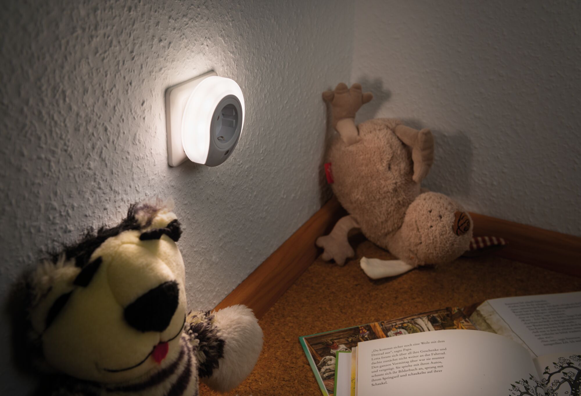 Weißes LED-Nachtlicht aus Kunststoff an Wand neben Kuscheltieren und offenem Buch im Kinderzimmer