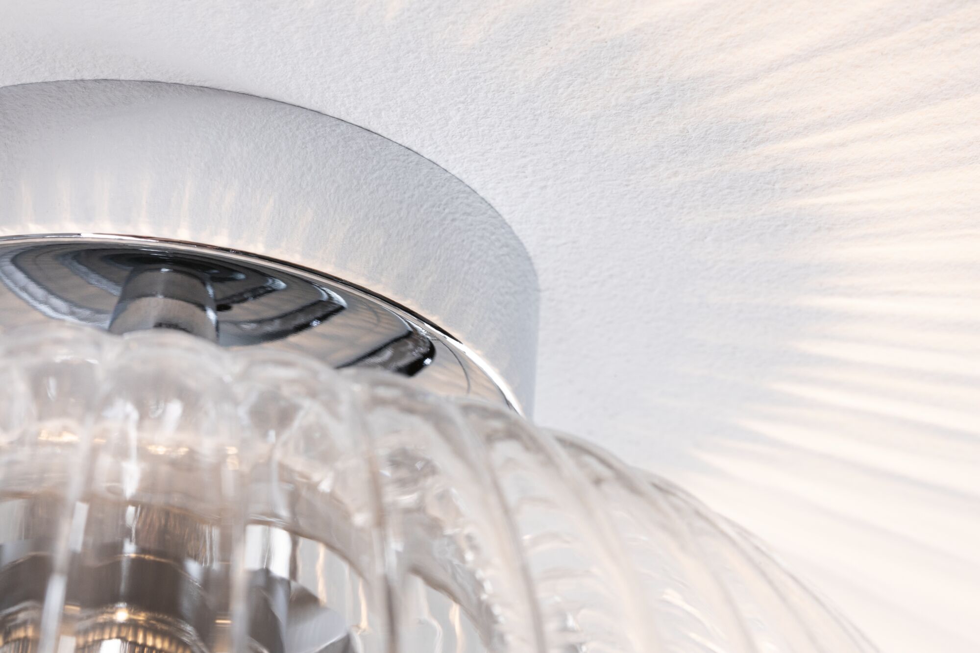 Nahaufnahme einer klaren Glasleuchte mit chromfarbenem Metallring und Lichtreflexionen an der Wand