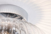 Nahaufnahme einer klaren Glasleuchte mit chromfarbenem Metallring und Lichtreflexionen an der Wand