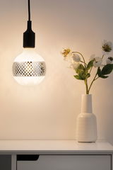 Ampoule suspendue avec câble noir et motif damier argenté à côté d'un vase blanc avec fleurs blanches sur table blanche