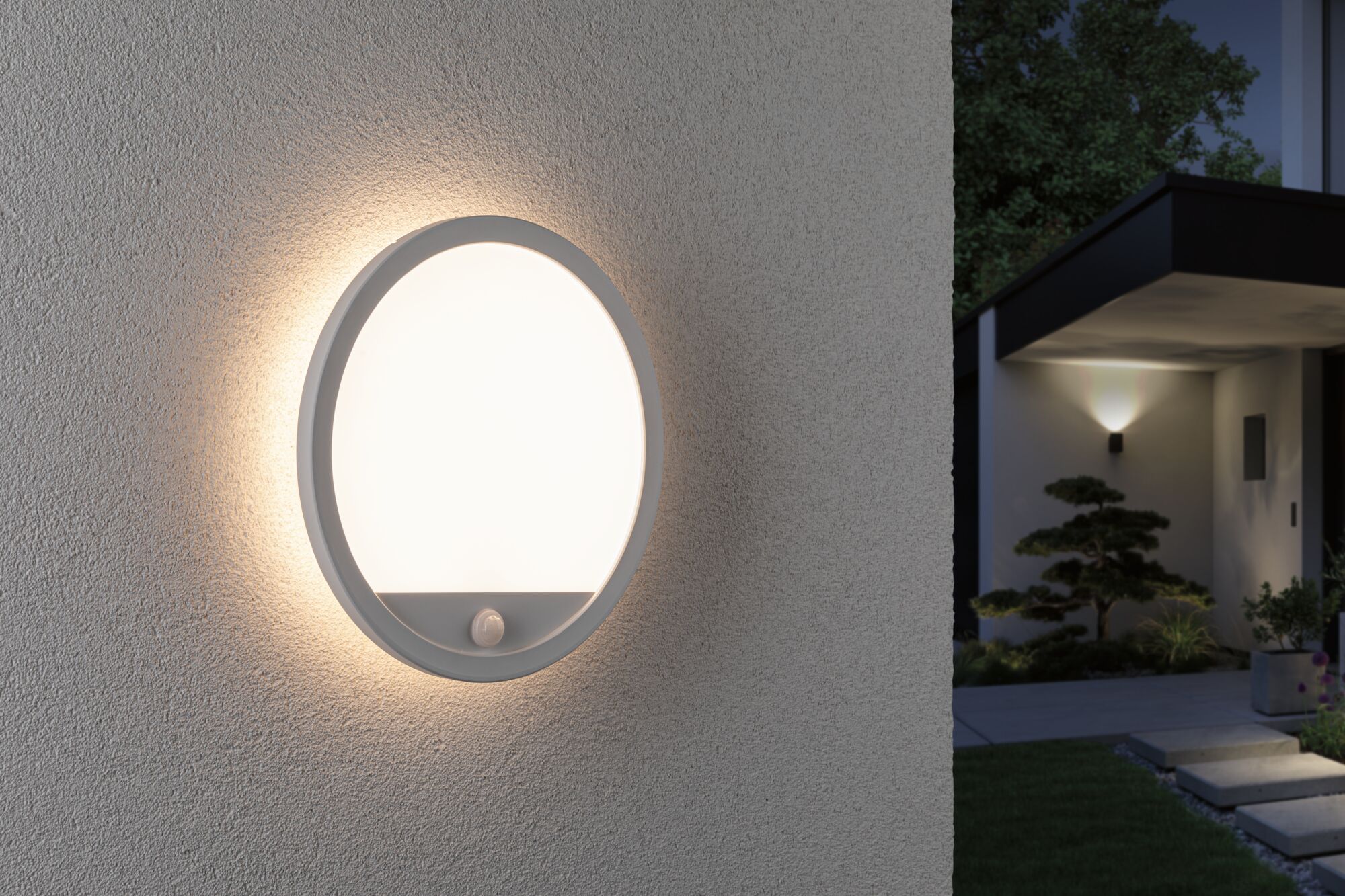 Round white outdoor wall light with motion sensor on grey wall illuminating entrance at night