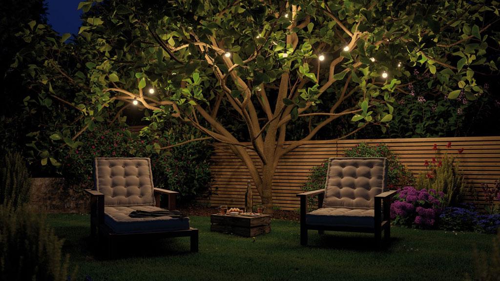 Éclairage de jardin chaleureux avec guirlandes LED blanc chaud sur arbre au-dessus de deux fauteuils rembourrés.