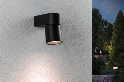 Black metal outdoor wall light with warm white illumination on white wall in garden setting.