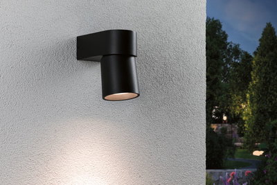 Schwarze Außenwandleuchte aus Metall mit warmweißem Licht an weißer Wand im Gartenbereich.