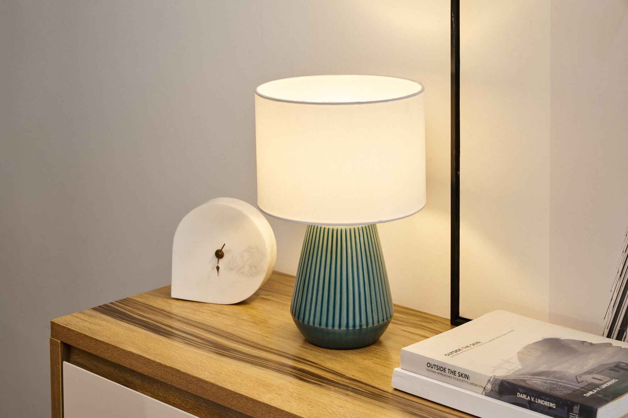 Tafellamp met blauwe keramische voet en witte stoffen kap op houten dressoir naast klok en boeken