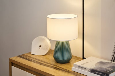 Tafellamp met blauwe keramische voet en witte stoffen kap op houten dressoir naast klok en boeken