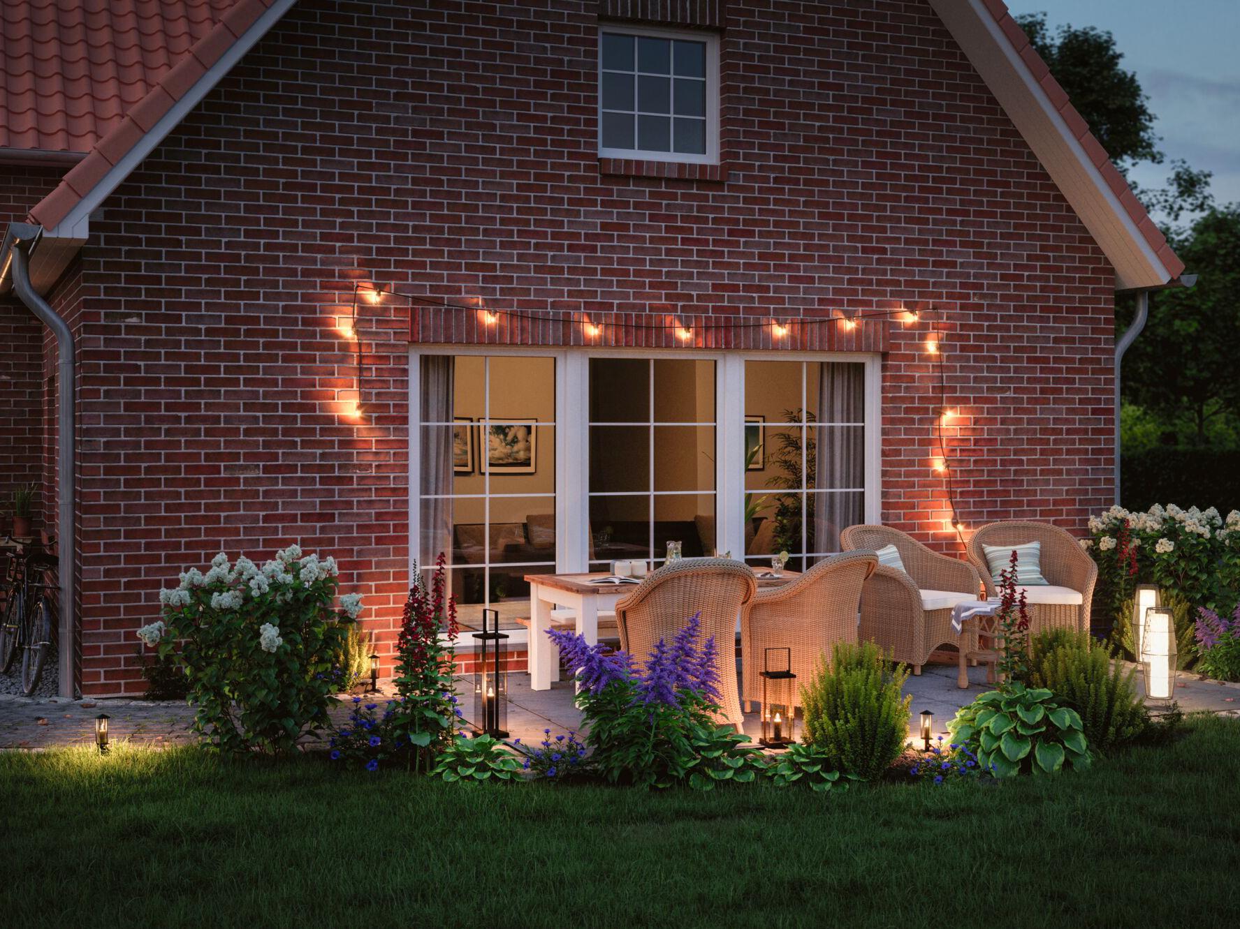 Gemütliche Terrasse mit Außenleuchten, warmem Licht und Gartenmöbeln aus Rattan vor rotem Backsteinhaus.