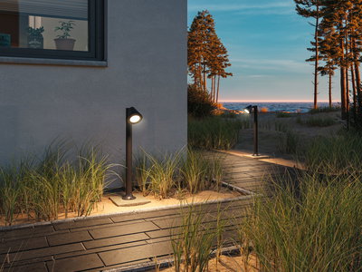Schwarze Außenwandleuchte aus Metall beleuchtet Weg mit Sand und Gras vor Haus bei Sonnenuntergang