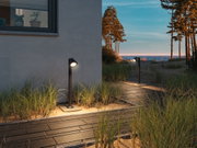 Black metal outdoor wall light illuminating sandy path with grass near house at sunset