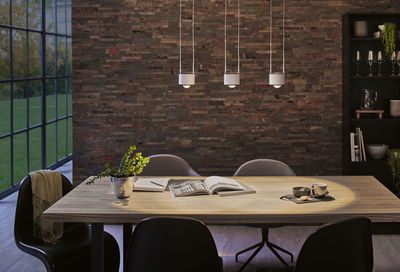 Moderner Essbereich mit hellem Holztisch, schwarzen Stühlen und drei weißen Pendelleuchten an Ziegelwand.