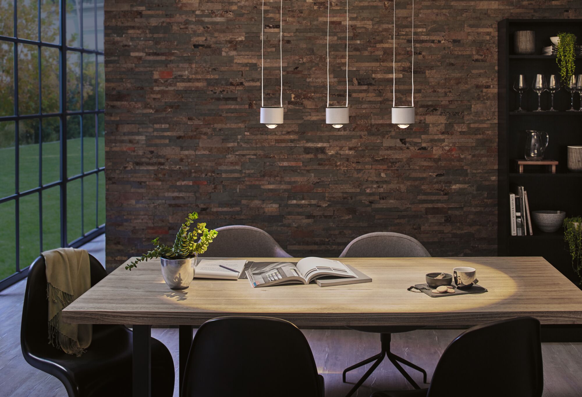 Salle à manger moderne avec table en bois clair, chaises noires et trois suspensions blanches sur mur en briques.
