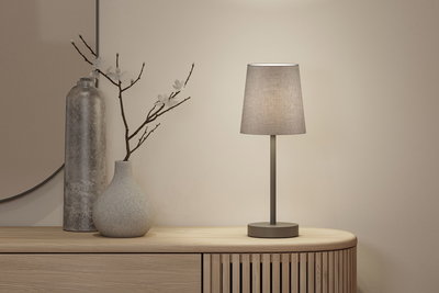 Moderne tafellamp met grijze stoffen kap en metalen voet op licht houten dressoir met decoratie.