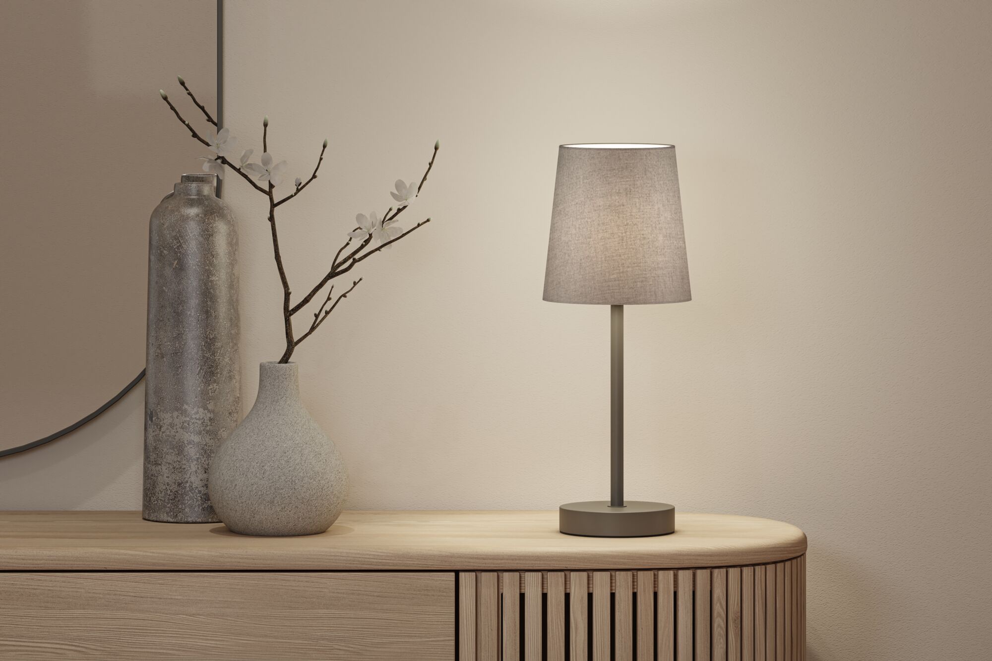Moderne Tischlampe mit grauem Stoffschirm und Metallfuß auf hellem Holzsideboard mit Deko.