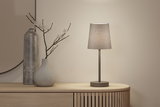 Moderne Tischlampe mit grauem Stoffschirm und Metallfuß auf hellem Holzsideboard mit Deko.